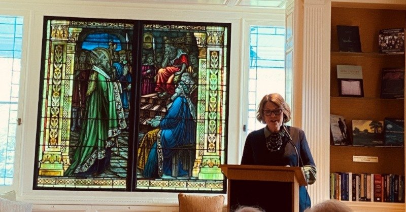 stained glass window with light behind it, in front of the window is a woman wearing glasses reading from a podium