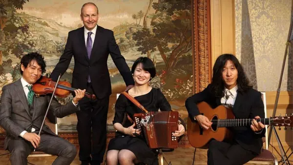 one man standing in a suit and tie behind three musicians seated with a violin, accordion and guitar, all arein front of an old painting