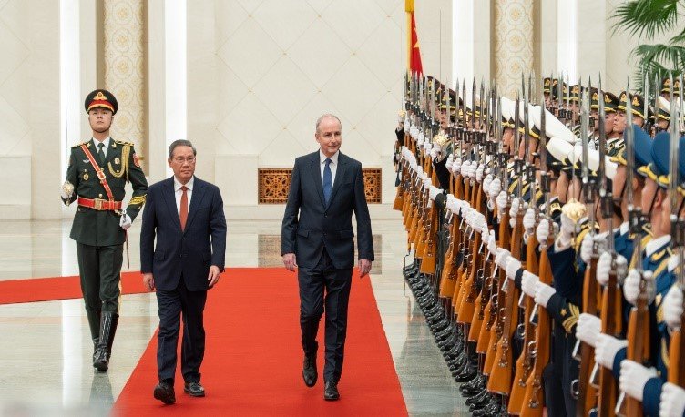 Two men walk on a red carpet with a row of military officers on the right. A military officer walks behind the men on the left.