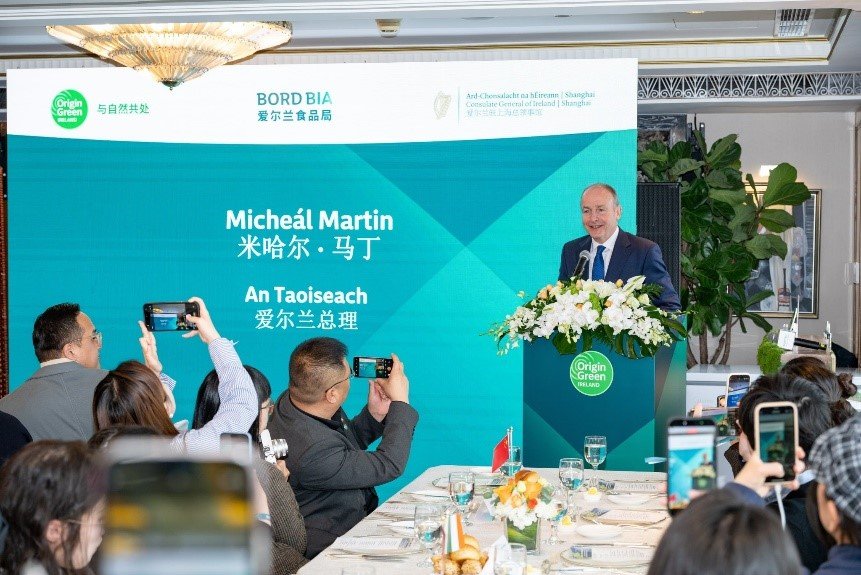 A man delivers a speech at a podiums. The audience is holding up phones to take photos. A backdrop to the left reads Micheál Martin, Taoiseach.