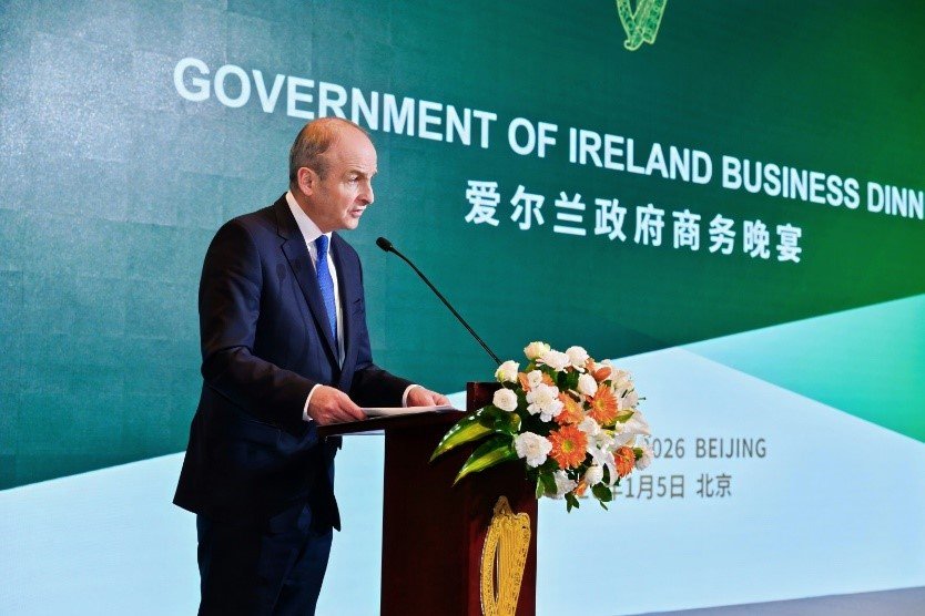 A man stands at a podium in front of a green backdrop which reads 'Government of Ireland business dinner.' There are white and orange flowers on the front of the podium.