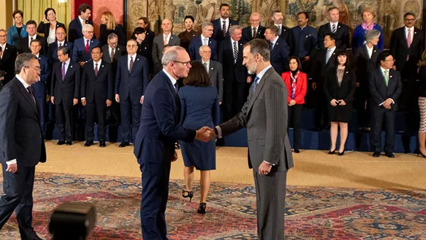 two men shaking hands on an old carpet with men and women in suits in three lines facing them