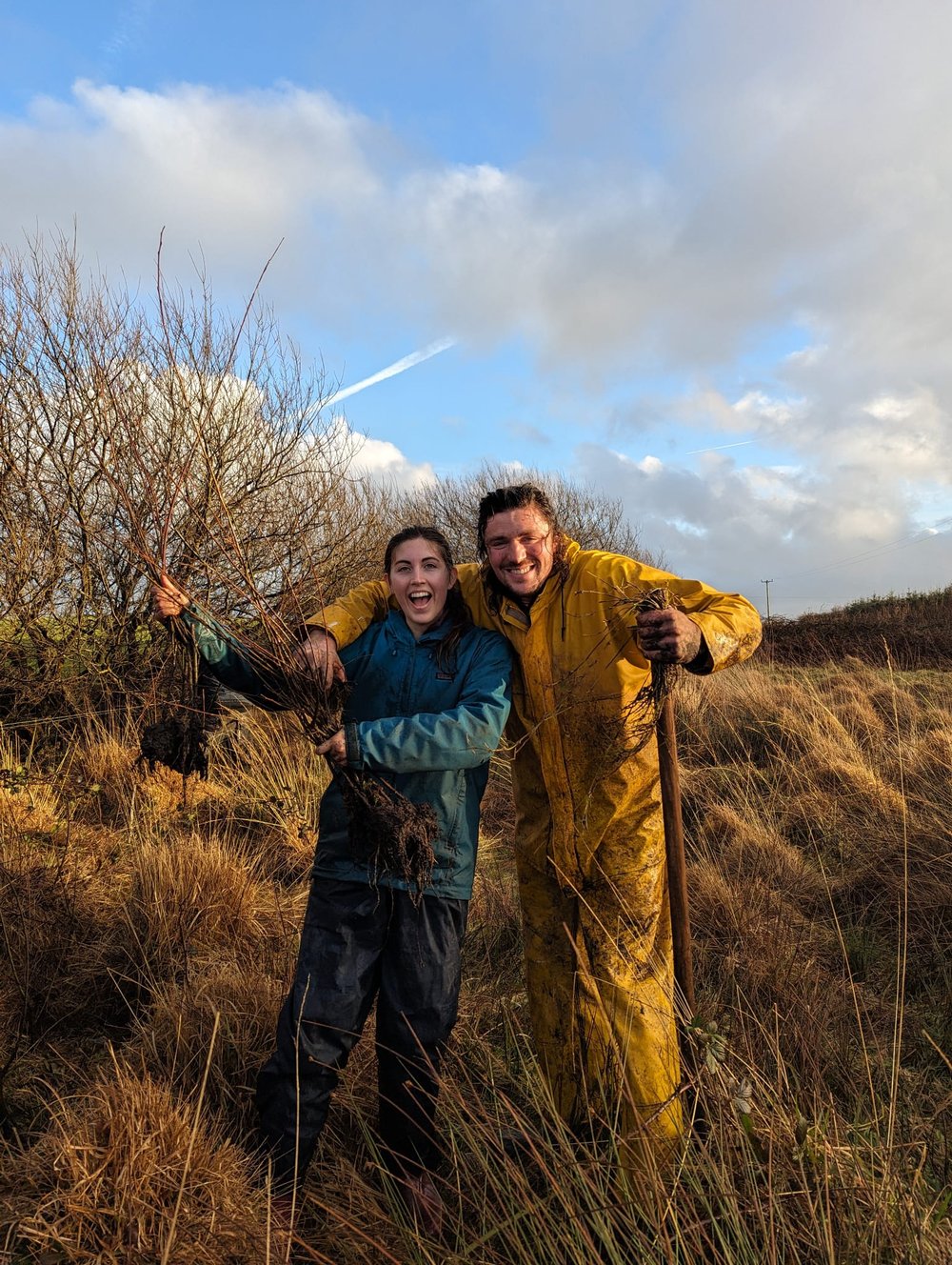 A young man and young woman dressed in warm clothes smile as they pose together in a field with young trees. The man holds a shovel.