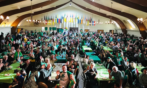 large banquet hall with hundreds of people wearing greeen sitting on long trestle tables with arched ceiling
