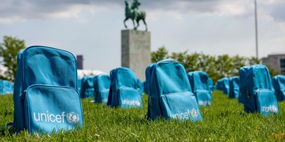 Sky blue backpacks with the UNICEF logo are laid out in rows as if they were gravestones. In the background is a statue of a horse and rider on a plinth and the UN flag flying on a flagpole.