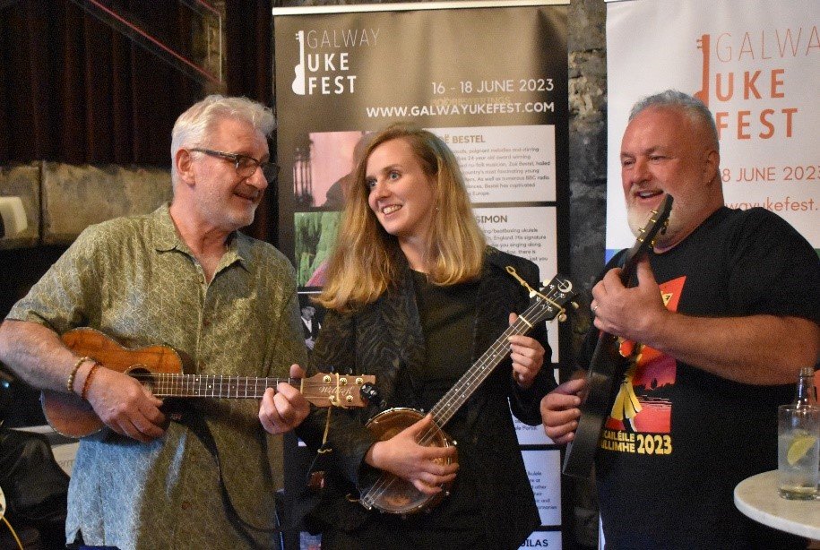Three musicians holding their instruments in front of a UkeFest sign