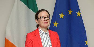 Ambassador Deirdre Ní Fhallúin headshot in front of flags