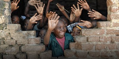Children waving through a gap in a brick wall.