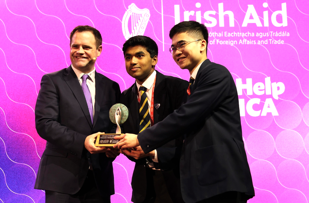 Man in suit presenting an award to two sudents and smiling in front of backdrop of screen