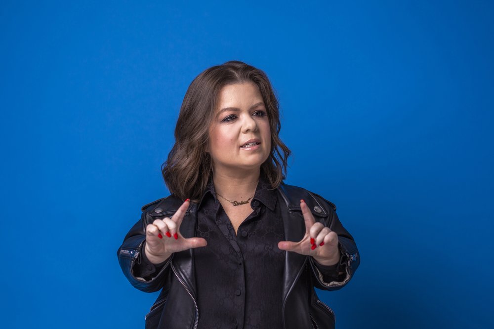 Sinéad Burke posing against a blue background, holding her hands up as if to frame a picture.