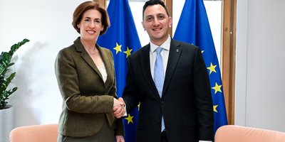 Minister Hildegarde Naughton shakes hands with Commissioner Glenn Micallef. Behind them, two EU flags are visible.