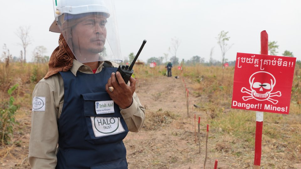A man wearing a helmet with a plastic face screen, and holding a walkie talkie, looks off camera. Beside him is a red sign with a skull and crossbones and the text: Danger! Mines!