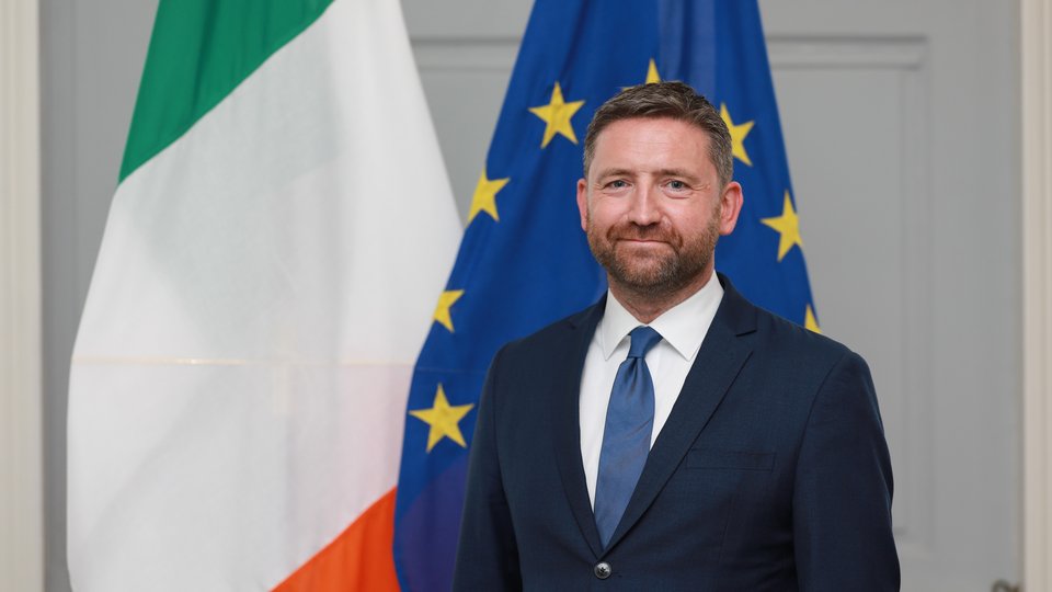 Headshot of Adrian Farrell standing in front of Irish and EU flags