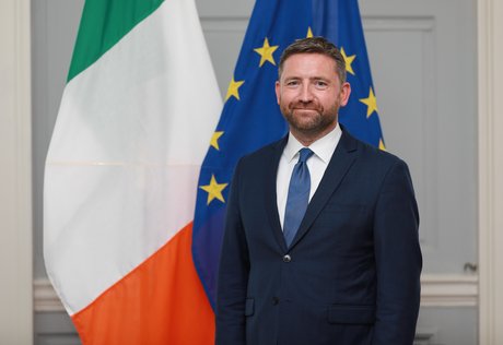 Headshot of Adrian Farrell standing in front of Irish and EU flags