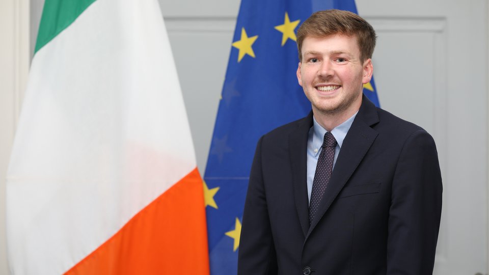 Headshot of a man in a suit standing in front of the Irish and EU flags.