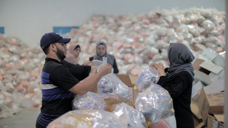 A man and woman tie transparent plastic bags with food items inside. Behind them are large piles of similar bags.