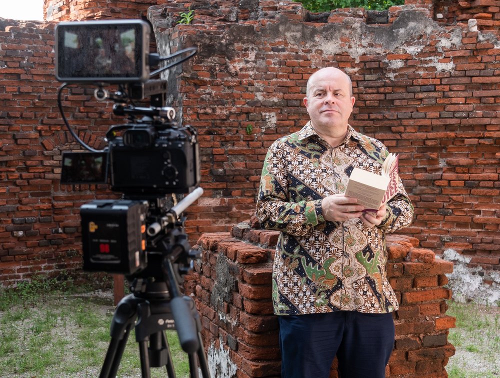 Ambassador reading from a book by a red brick wall in front of a camera