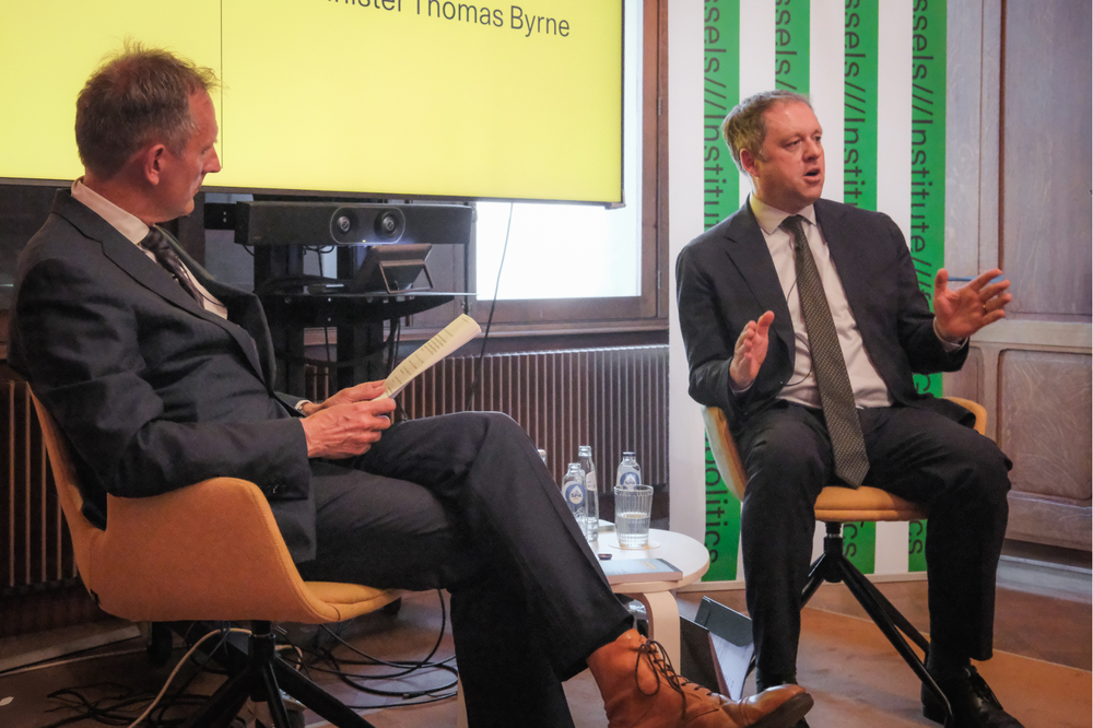 Minister Byrne sitting on stage, speaking at a think tank engagement. To his left, is a low table upon which bottles of water and a half-filled glass are visible. The event moderator sits to his left also, holding papers.