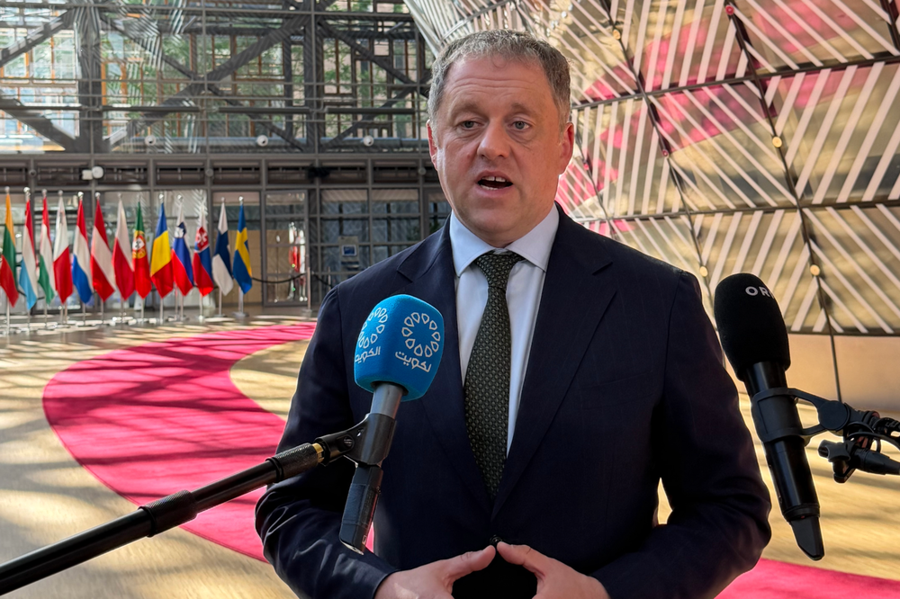 Minister Byrne stands in the Council forum, speaking to media ahead of the General Affairs Council. The forum, Member State flags, and a red carpet are visible behind him. Two microphones are placed in front of him.