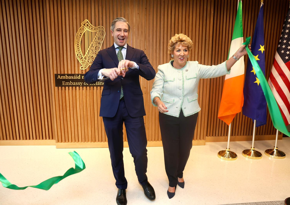Tánaiste Simon Harris and Ambassador Byrne Nason cut green ribbon in front of wood background with Irish, EU, and American flags.